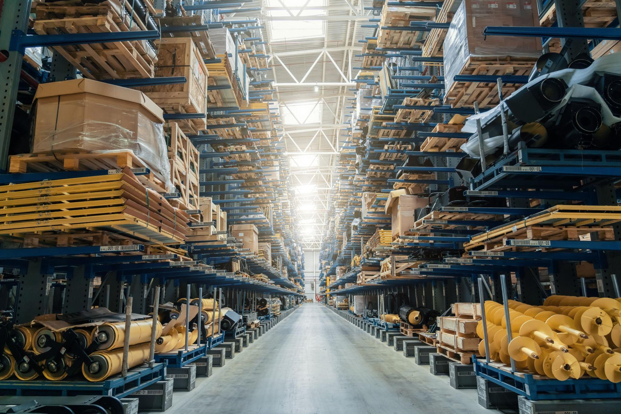 A long indoor perspective of wide warehouse storage aisles featuring high industrial shelves organized with pallets, crates, and manufacturing materials.